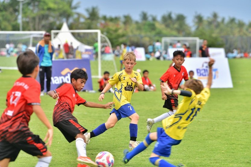Young players in yellow and red chasing the ball at BALI7s